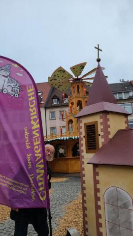 Rechts die Wagenkirche mit Turm, links eine Beachflag mit Aufschrift "CitykWagenKirche - schnell mal Kirche". In Hintergrund die große Weinachtspyramide des Weihnachtsmarkts. Lorenz Hummel lugt lachend hinter der Fahne vor.