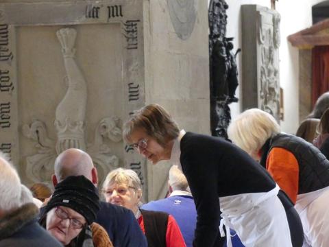 Regionalbischöfin Gisela Bornowski mit Vesperkirche-Schürze beugt sich als Gastgeberin freundlich zu Gästen am Tisch, die in der Kirche sitzen.