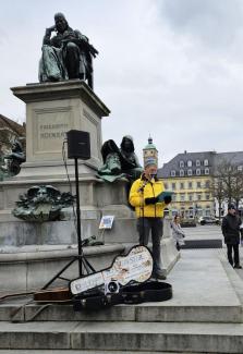 Christian Schäflein steht in gelber Jacke am Rückertdenkmal an einem Mikrofon. Im Hintergrund der Turm der St. Johanniskirche. Vorne liegt ein offener Gitarrenkoffer des Musikers Erik Stenzel.